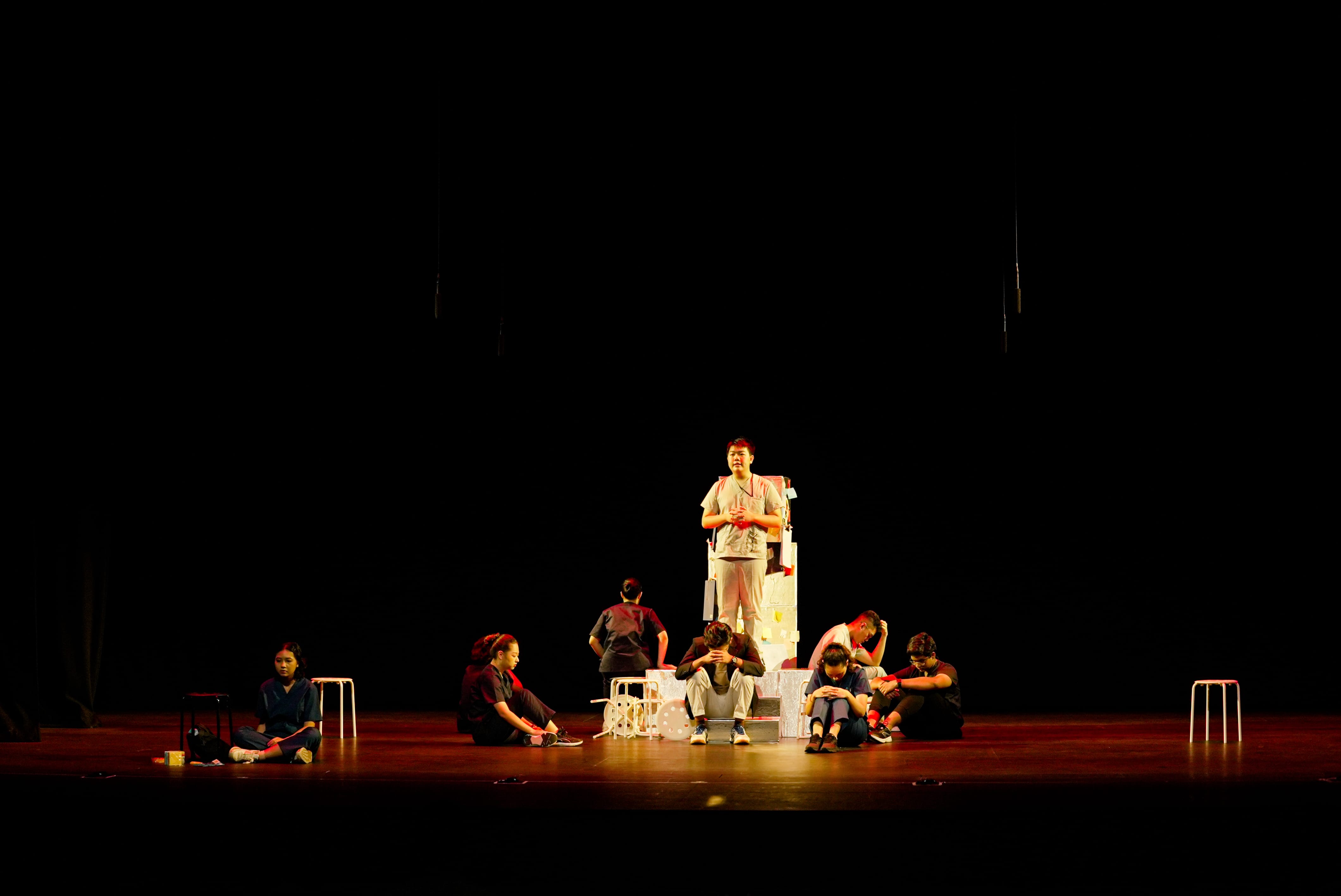 A lone student stands elevated on a white prop structure at centre stage while eight other students sit or crouch around them in poses of distress, lit by a warm amber spotlight.
